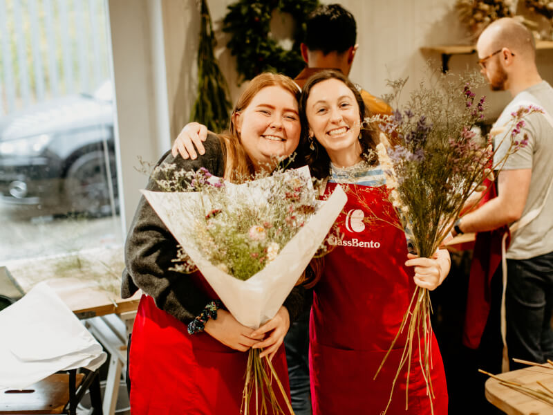 Two young women in red aprons smiling while holding bouquets of dried flowers